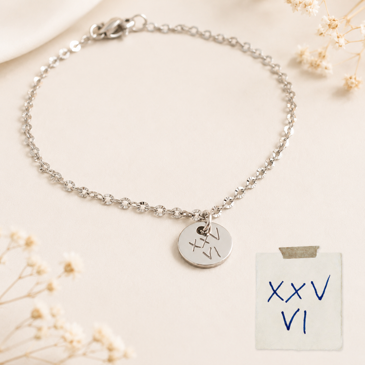 Gold chain bracelet with a round engraved heart charm, laid on beige fabric with dried flowers
