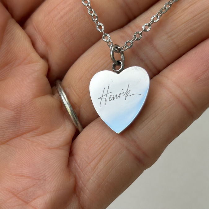 Silver heart necklace engraved with a handwritten signature, held in the palm