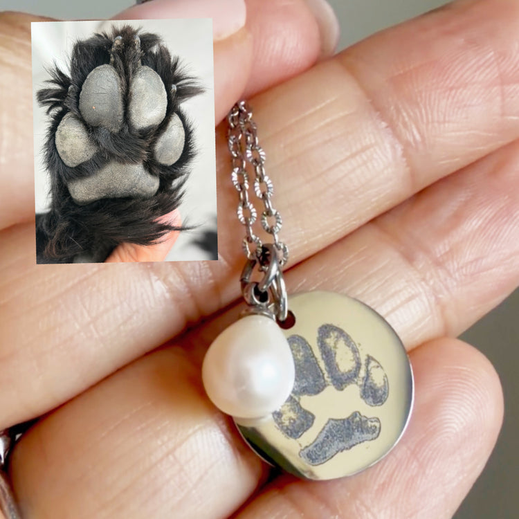 Hand holding a silver pet paw print necklace with a pearl charm, next to the original paw print