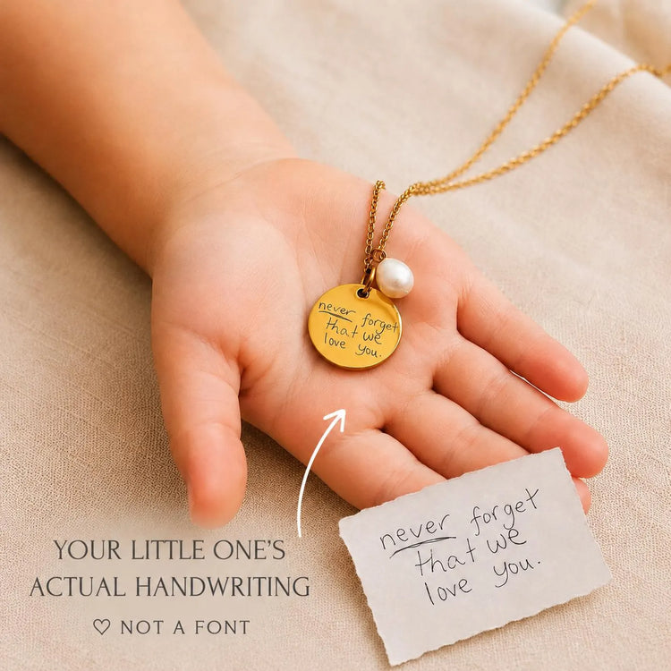A child's hand holding a custom engraved gold necklace with a round engraved gold pendant and a small white pearl bead. 