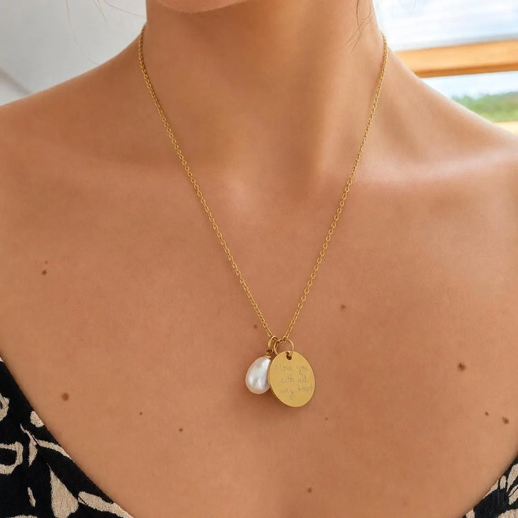 A gold chain necklace with two pendants worn on a model's neck: a round engraved gold disc and a white pearl-like drop, shown against bare skin.