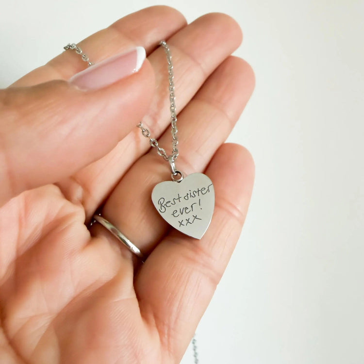 Hand holding a small silver heart necklace engraved with handwritten 'Best sister ever'