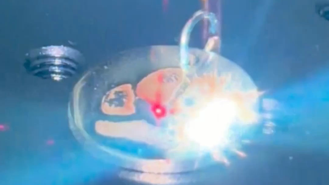 A close-up of a small clear oval container or dish with a bright white light flare in the center, surrounded by blue lighting and dark reflective surfaces.