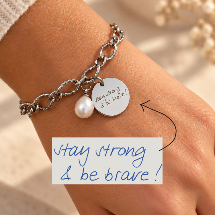 Silver chain bracelet on the wrist engraved with handwritten 'stay strong & be brave', beside the original writing