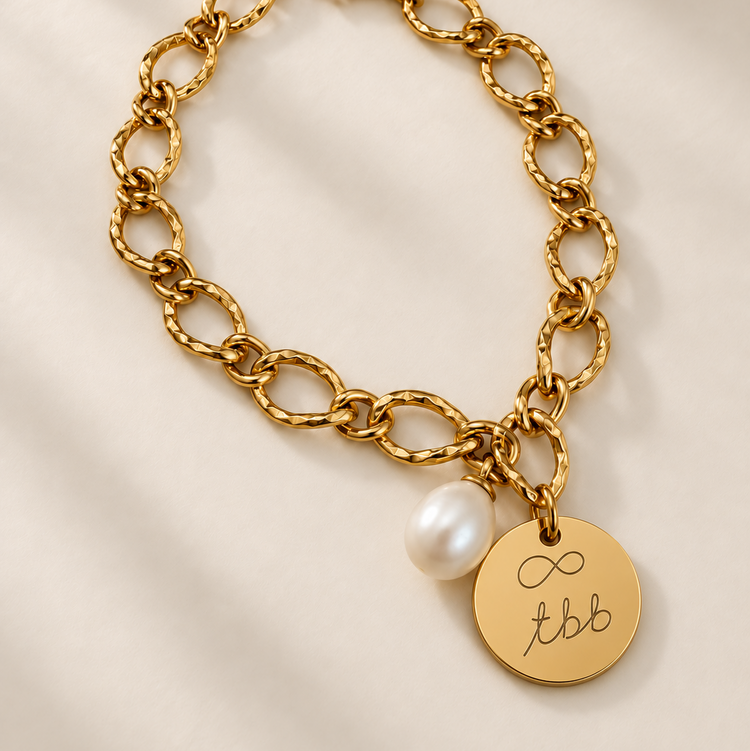 Gold chain bracelet with a disc charm engraved with a handwritten infinity symbol and initials, plus a pearl